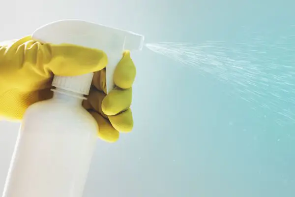 Pessoa de luva plástica amarela utiliza álcool spray para limpar ambientes