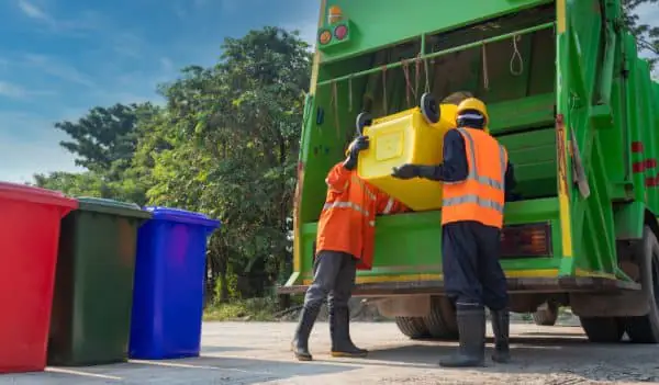 Três contêineres de lixo nas cores vermelho, preto e azul ao lado de um caminhão de lixo verde, onde dois trabalhadores vestindo EPIs descartam o conteúdo do contêiner de lixo amaralo. 
