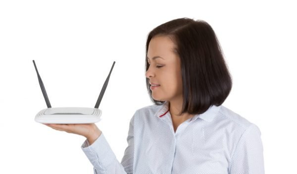 Mulher de camisa branca, cabelo escuro na altura dos ombros segura na mão direita um roteador de internet branco com antenas pretas.