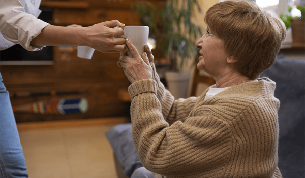 Mulher idosa com um casaco de lã bege recebendo uma xícara de café em uma sala de espera