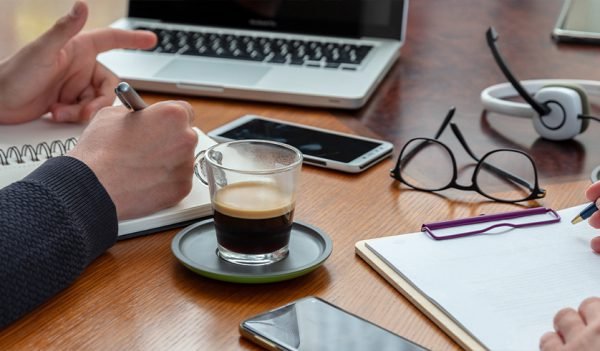 Mesa de trabalho com uma xícara de café, celulares, um computador e duas pessoas fazendo anotações em agendas
