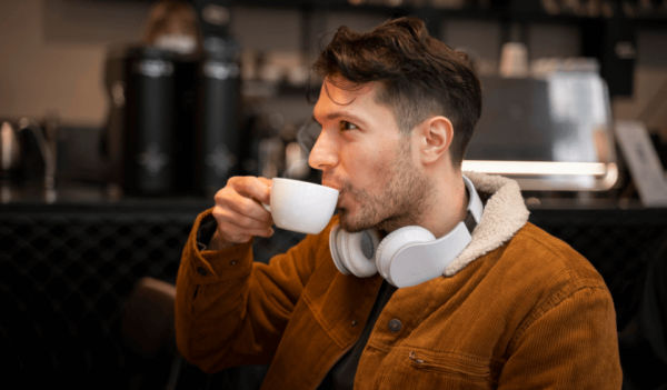 Homem jovem com casacos e fones de ouvidos bebe café em uma cafeteria