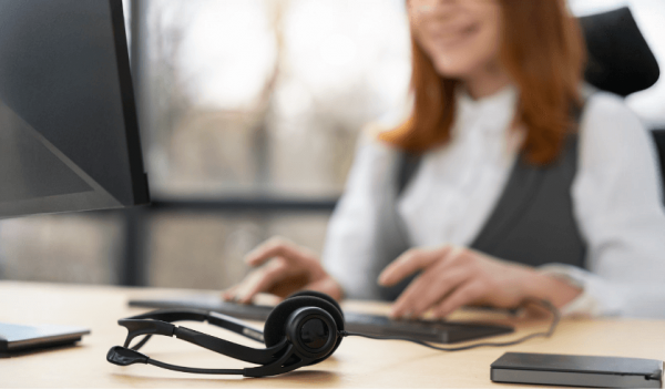 Headset posicionado na mesa de uma profissional que digita no teclado do seu computador.